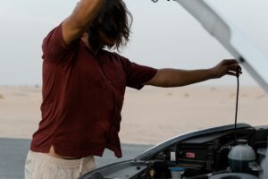 A man examines the engine of a car on a deserted road, highlighting automotive maintenance.